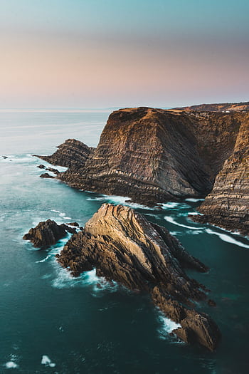 A breathtaking view of rugged cliffs and ocean waves in Beja District, Portugal during a serene sunset. HD phone wallpaper