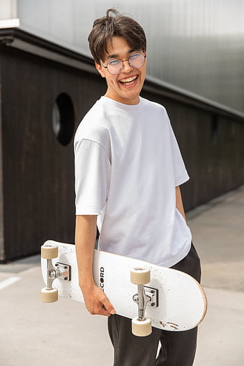 Happy young man with glasses holding a skateboard at a skatepark, laughing with joy. HD phone wallpaper
