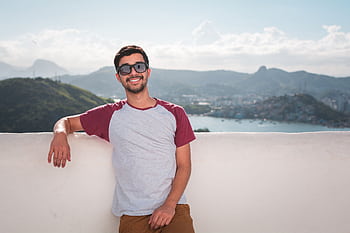 Young man in sunglasses leaning on a balcony with a scenic city view in the background. HD wallpaper