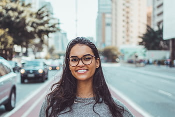A young woman with glasses smiling on a city street, embracing urban lifestyle. HD wallpaper
