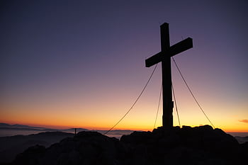A dramatic cross stands silhouetted against a colorful sunset on a mountain peak, symbolizing faith and peace. HD wallpaper