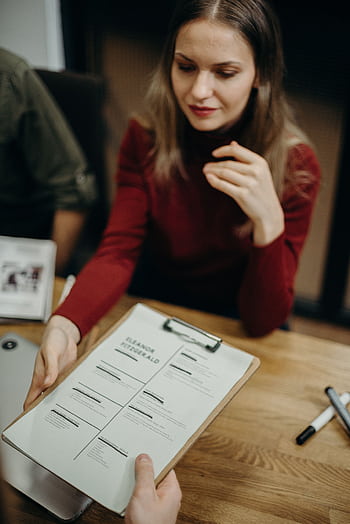 Woman sitting at a desk holding a clipboard and reviewing a resume during a job interview. HD phone wallpaper