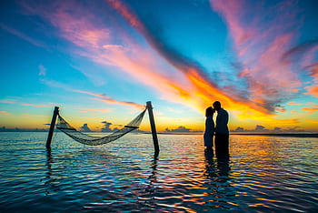 A romantic couple shares a kiss at sunset with vibrant skies over the Maldives beach. HD wallpaper