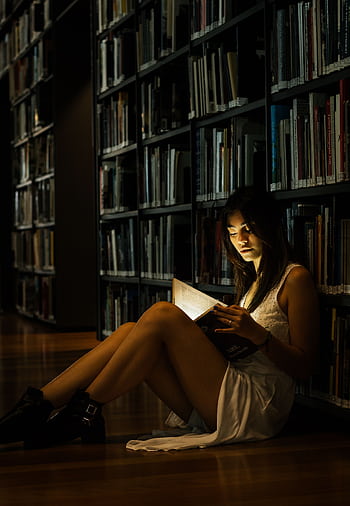 A woman reads a glowing book on the floor of a dimly lit library, creating a magical atmosphere. HD phone wallpaper