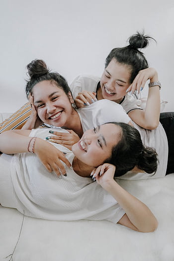 Three young women laughing and embracing each other indoors, showcasing friendship and happiness. HD phone wallpaper