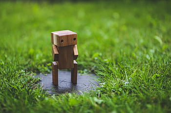 A small wooden toy robot stands on grass, surrounded by vibrant greenery. HD wallpaper