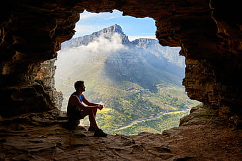 A man sitting in a cave overlooking a majestic mountain landscape under daylight. HD wallpaper