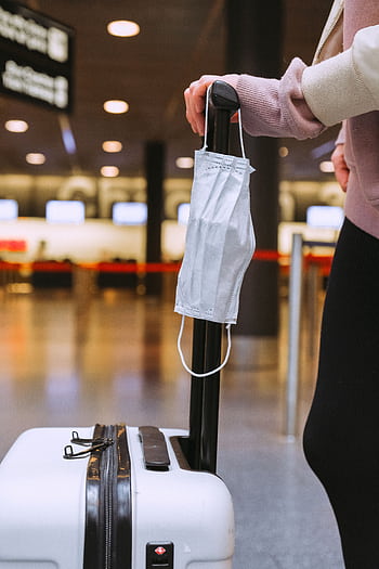 Person standing with luggage and face mask hanging, symbolizing pandemic travel. HD phone wallpaper