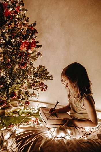 A young girl writes in a notebook by a Christmas tree adorned with decorations and lights. HD phone wallpaper