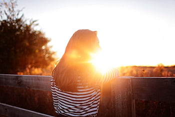 A woman leans against a wooden fence, basking in the warm glow of a sunset on a rural farm. HD wallpaper