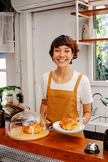 Charming waitress in apron serving croissant with a smile in a cozy café setting. HD phone wallpaper