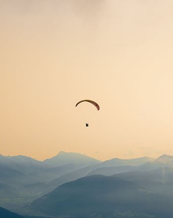 A paraglider soars gracefully over the misty, layered mountains at sunset, capturing serene adventure. HD phone wallpaper