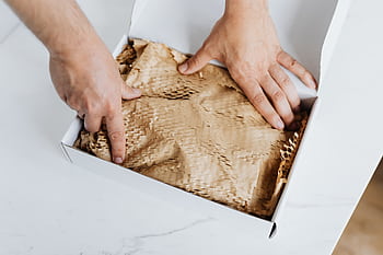 Close-up of hands packing an eco-friendly cardboard box with protective paper, showcasing sustainable packaging. HD wallpaper