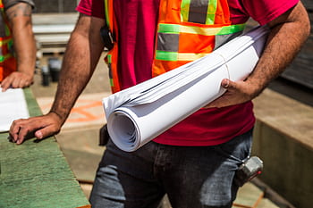 Construction workers in safety vests holding blueprints at a site during the day. HD wallpaper