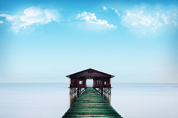 Tranquil scene of a pier leading to a cottage over a calm ocean under a bright blue sky. HD wallpaper