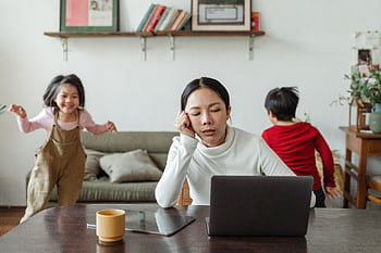A tired mother working on a laptop while her children play around indoors, highlighting remote work challenges. HD wallpaper