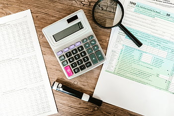 Top view of tax forms, a calculator, and a magnifying glass on a wooden surface, ideal for accounting themes. HD wallpaper