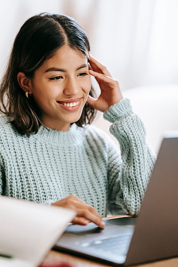 A young woman smiling as she uses a laptop in a cozy indoor setting, creating a comfortable workspace atmosphere. HD phone wallpaper