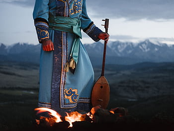 A traditional musician in ornate attire plays an instrument by a bonfire against a mountainous backdrop at dusk. HD wallpaper