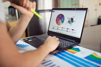 Person analyzing financial charts and graphs on a laptop with colorful documents, showcasing market analysis. HD wallpaper