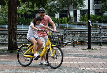 Father teaches daughter to ride a yellow bicycle in a Singapore park. Bonding moment. HD wallpaper