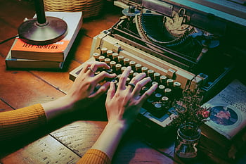 A cozy scene with a vintage typewriter and hands typing indoors beside books and flowers. HD wallpaper