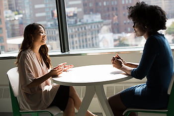 Two professional women having a discussion at a desk in a bright office setting. HD wallpaper