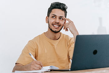 Smiling young man in yellow shirt thoughtfully planning project while using laptop indoors. HD wallpaper