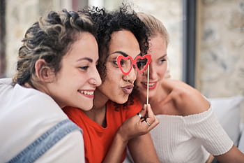 Three women enjoying a playful moment with heart-shaped glasses, symbolizing friendship and fun. HD wallpaper