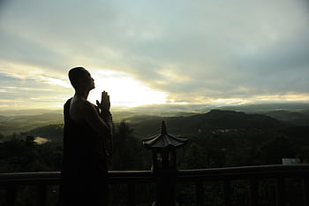 A monk in silhouette prays peacefully at sunrise with mountains in the background. HD wallpaper