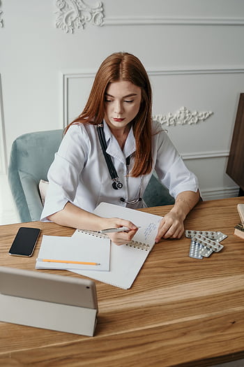 A female doctor writes notes at her desk in a clinical setting, focusing on healthcare tasks. HD phone wallpaper