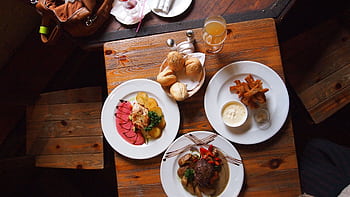 Aerial view of a rustic wooden table set with gourmet dishes, bread, and a drink, creating a warm dining atmosphere. HD wallpaper