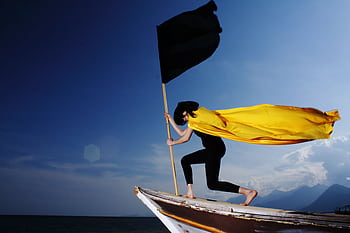 A woman in a bold pose with a black flag and yellow cape on a boat by the sea under blue skies. HD wallpaper