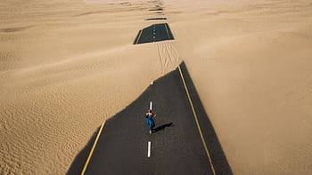 A lone woman walking on a sand-covered road in Dubai's desert, captured from above. HD wallpaper
