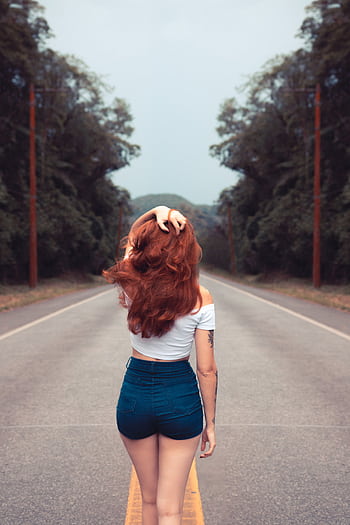 Red-haired woman in jean shorts walking down a forest road, showcasing style and dom. HD phone wallpaper