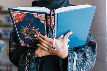 A Muslim woman in traditional attire reading the Quran indoors, holding prayer beads in a peaceful setting. HD wallpaper