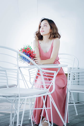 A woman in a pink dress holding colorful flowers while sitting indoors on white chairs. HD phone wallpaper