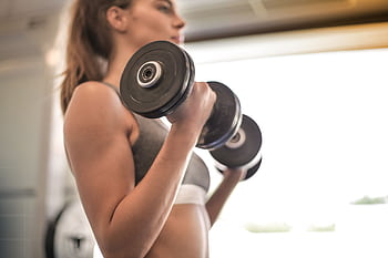 Focused woman lifting dumbbells in a bright gym, emphasizing strength and fitness. HD wallpaper