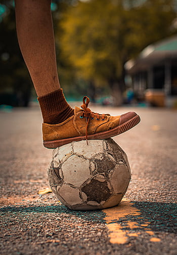 An aged soccer ball under a foot in casual footwear on an outdoor surface. HD phone wallpaper