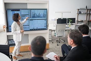 A woman presents data on a screen to a group of colleagues in an open office environment. HD wallpaper
