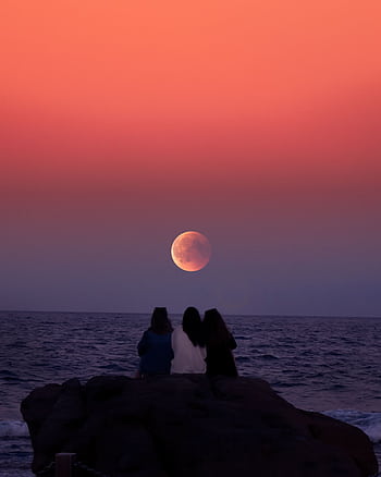 Three friends silhouetted against a vibrant twilight sky, gazing at the moonrise over the ocean. HD phone wallpaper