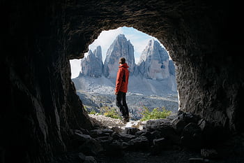 Man in red jacket standing in cave entrance, viewing Tre Cime di Lavaredo in Italy. HD wallpaper