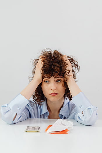A worried woman with curly hair sits with her hands on her head surrounded by bills. HD phone wallpaper