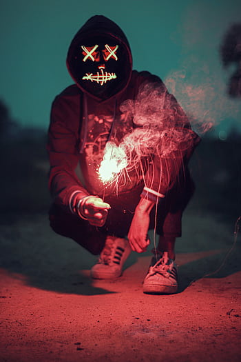 Creepy figure with neon mask crouched outdoors, holding a sparkling light in the dark. HD phone wallpaper