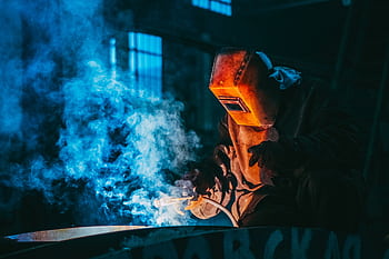 A welder in a workshop using safety gear to weld metal, surrounded by blue smoke and atmospheric lighting. HD wallpaper