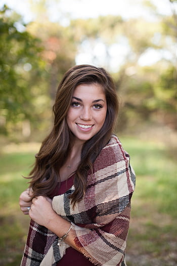 A woman smiling outdoors with a cozy plaid shawl, surrounded by nature in a sunny park. HD phone wallpaper
