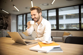 Man enjoying a coffee break while working in a modern office with laptop and books. HD wallpaper