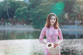 Woman in traditional pink Ao Dai dress smiling by a tranquil lake in daytime outdoor setting. HD wallpaper