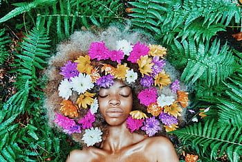 A serene portrait of a woman with flowers in her afro, surrounded by lush green ferns. HD wallpaper