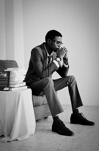 A thoughtful man in formal wear sits indoors, surrounded by books, conveying a pensive mood. HD phone wallpaper